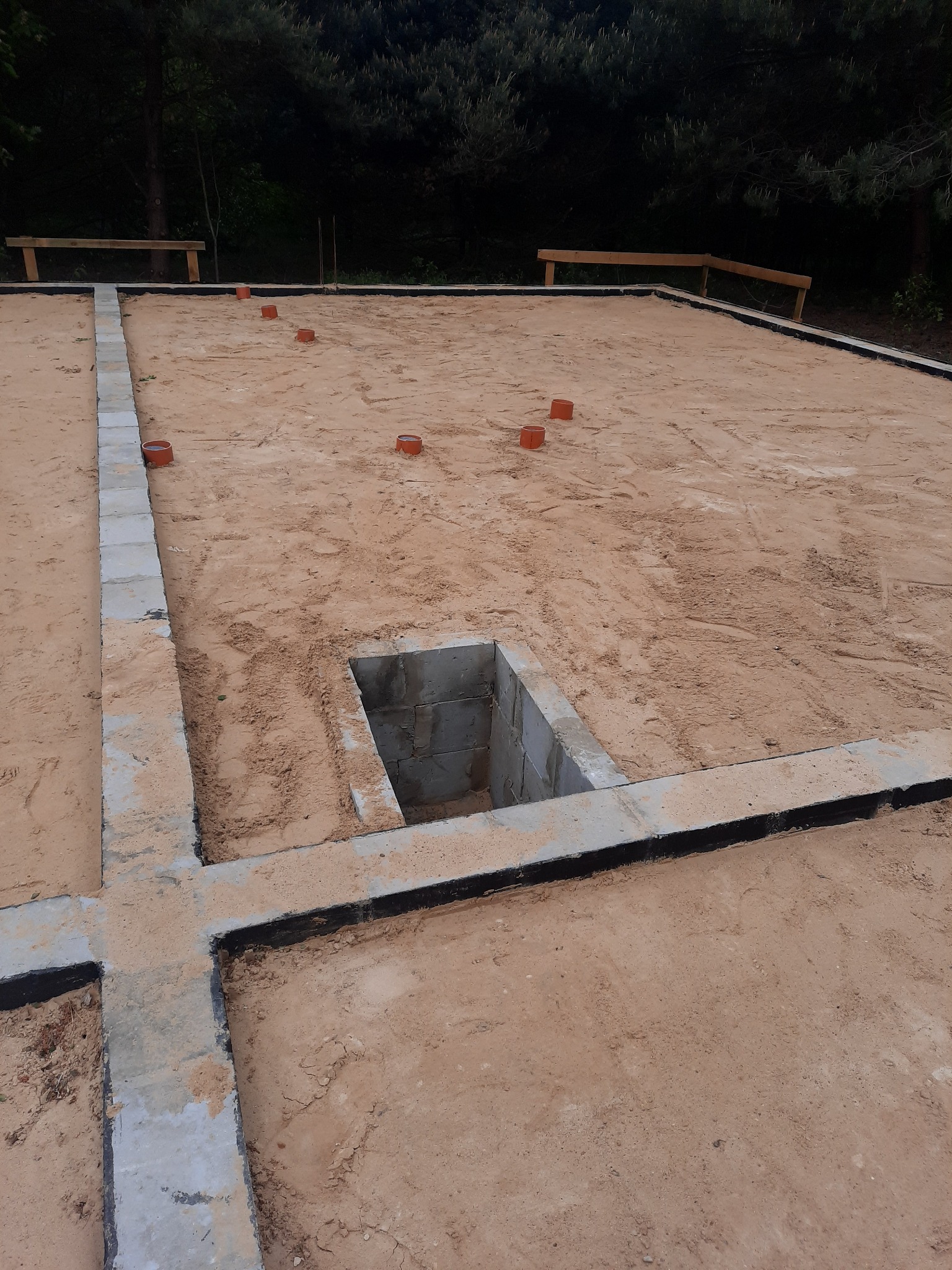 Zarys fundamentów domu z betonowych bloczków, widoczny wykop na instalacje podposadzkowe, teren wyrównany piaskiem, w tle zieleń.