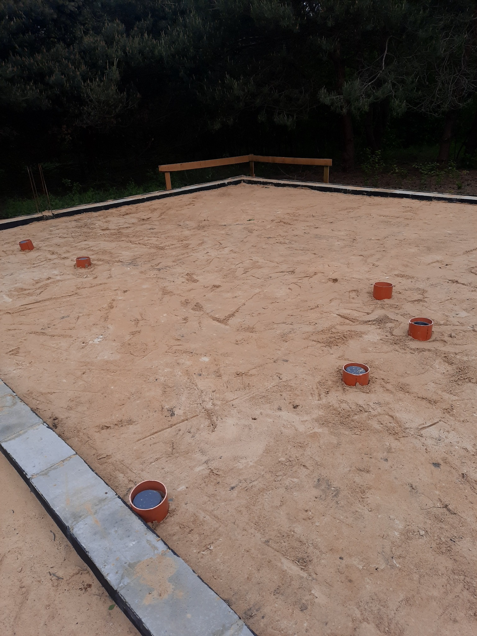 Przygotowany teren pod fundament z betonowymi obrzeżami i wystającymi rurami kanalizacyjnymi, otoczony drzewami.