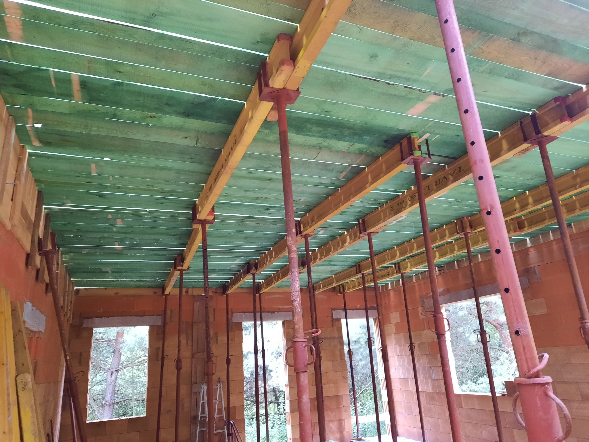 Wnętrze budowanego domu z czerwonej cegły ceramicznej, z widocznym deskowaniem stropu podpartym metalowymi stemplami w kolorze czerwonym i żółtym, widok przez okna na drzewa.