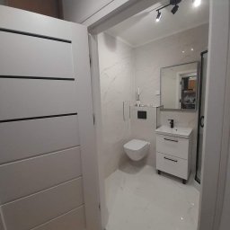 Idealbud - Nowoczesna łazienka z białymi marmurkowymi płytkami, wiszącą toaletą, szafką z umywalką i czarnym oświetleniem punktowym, widziana zza uchylonych drzwi.