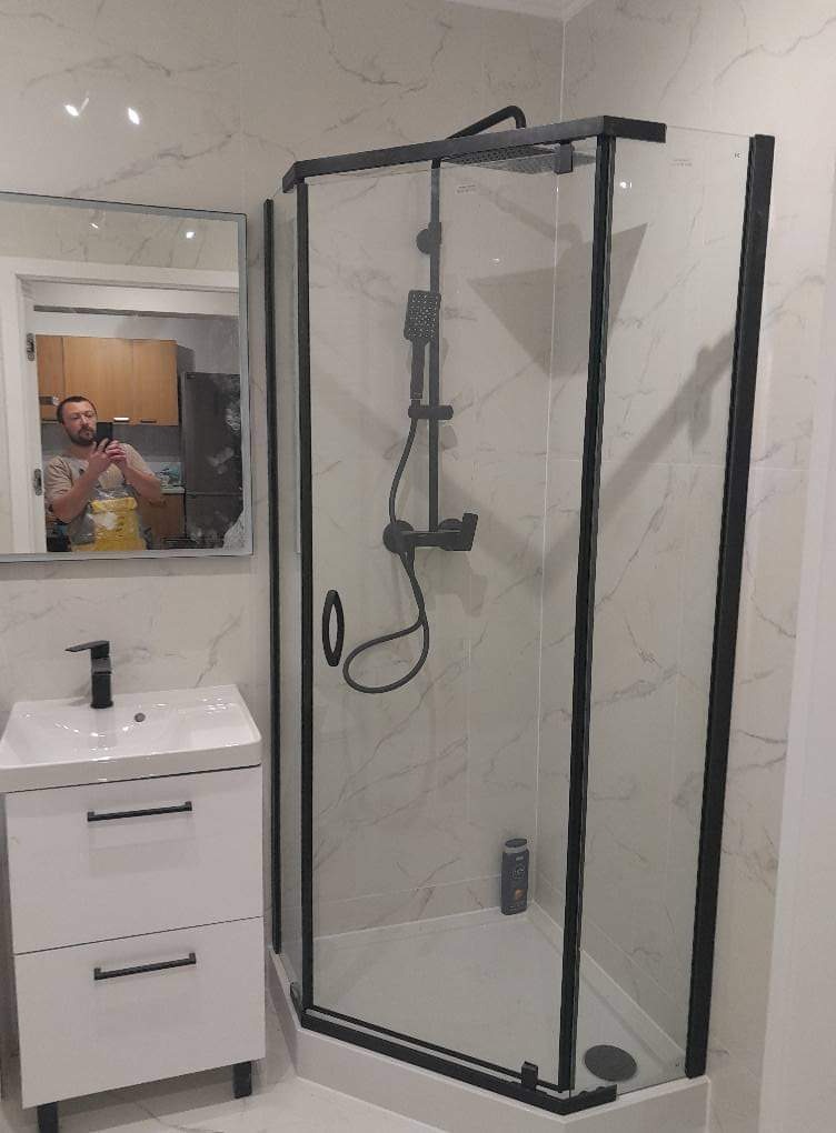 Nowoczesna kabina prysznicowa z czarnymi profilami i deszczownicą, biała umywalka z szafką w tle, odbicie osoby robiącej zdjęcie w lustrze.