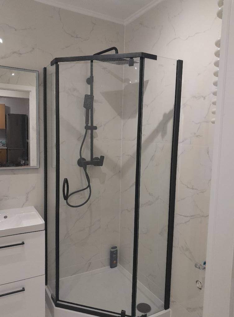 Nowoczesna kabina prysznicowa z czarnymi profilami na tle marmurowych płytek, widoczna umywalka z szafką w rogu pomieszczenia.