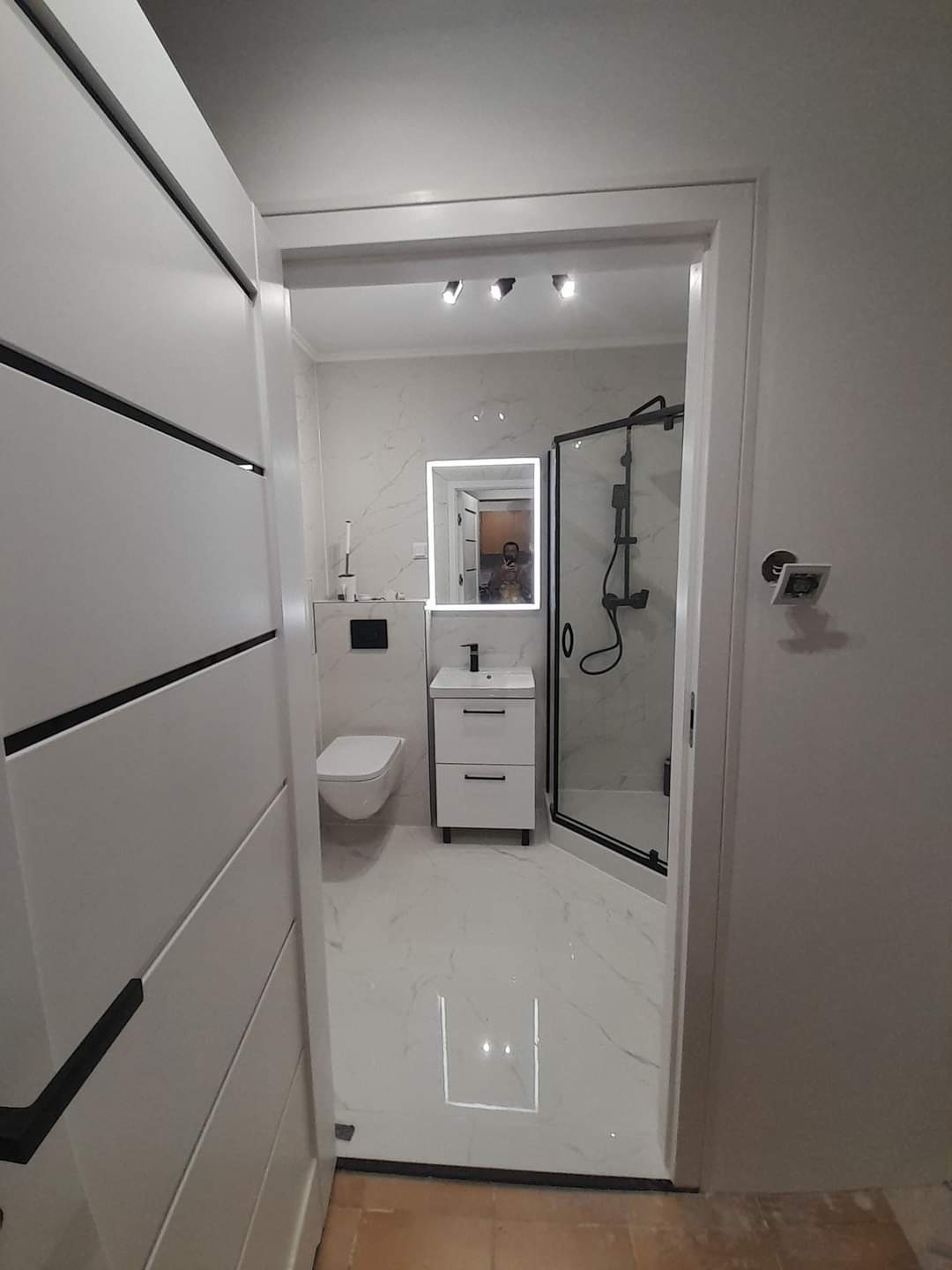 Nowoczesna łazienka z kabiną prysznicową z czarnymi ramami, białą armaturą i podświetlanym lustrem, widoczna przez otwarte drzwi z dekoracyjnymi frezami.