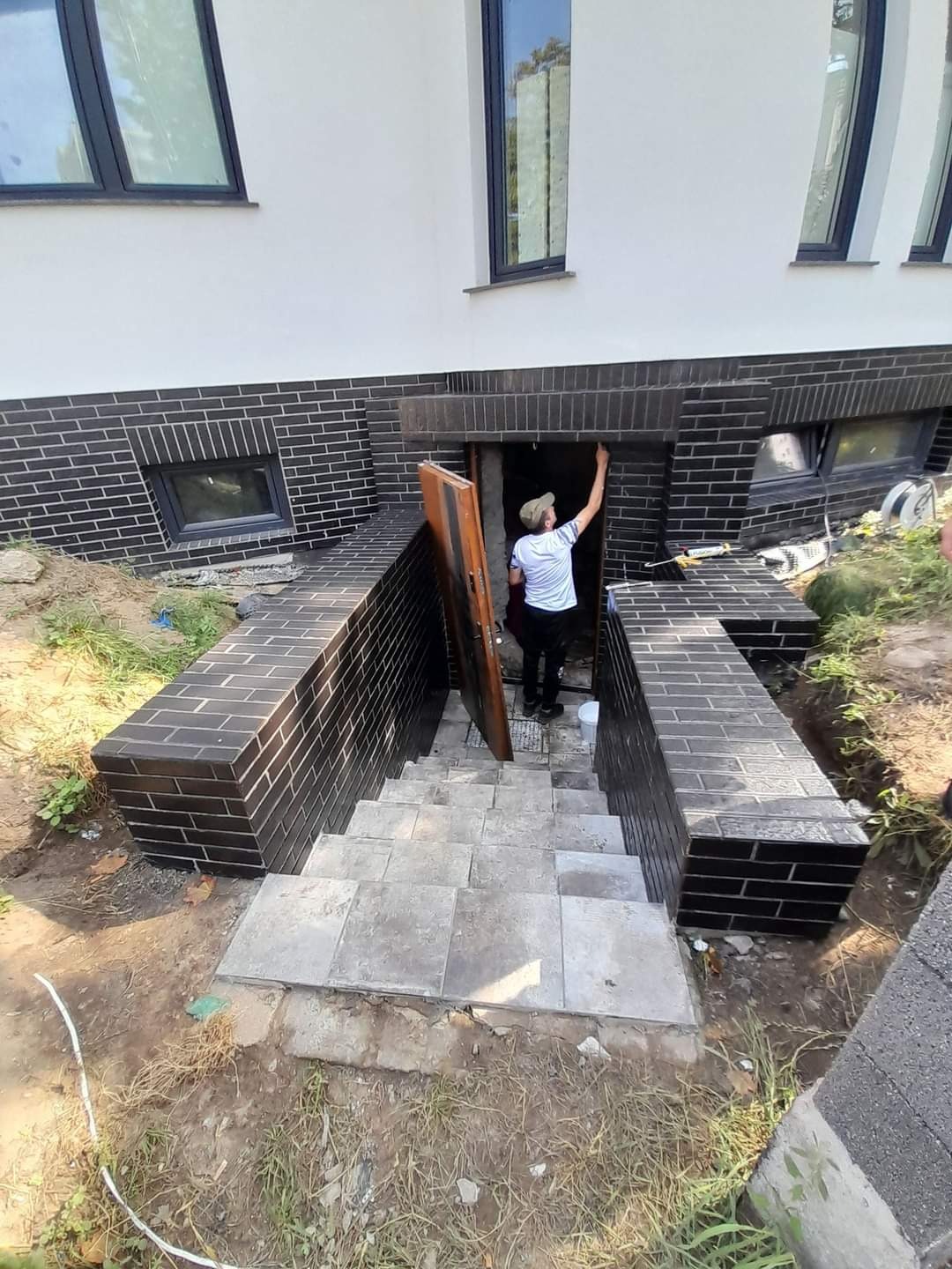 Pracownik maluje ciemną farbą wnękę wejścia do piwnicy z ceglanymi ścianami i schodami z szarych płyt betonowych, widoczne świeże ślady farby na cegłach.