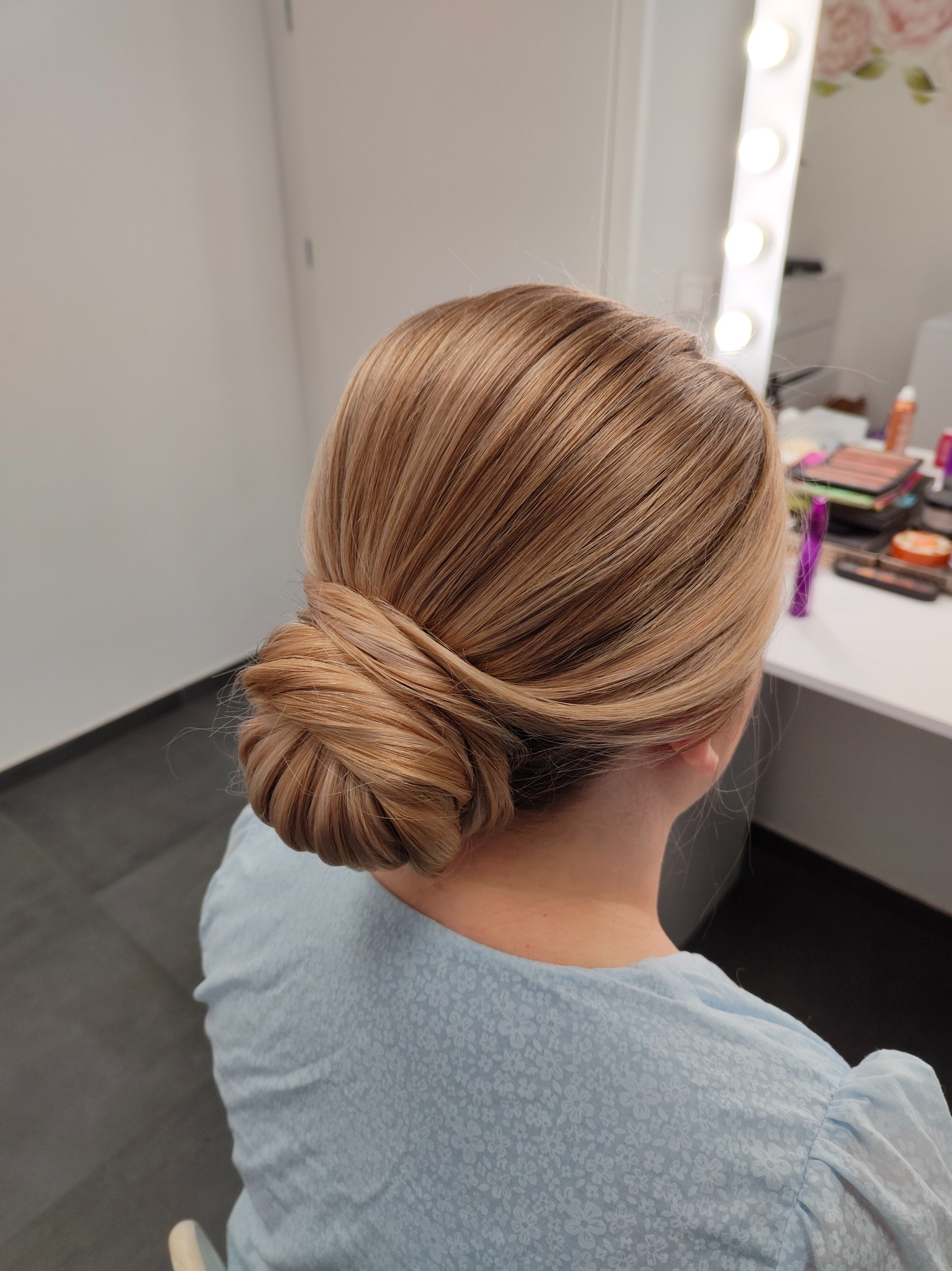 Eleganckie upięcie włosów blond w kok na karku, wykonane w salonie w Oławie, widok z tyłu, w tle toaletka z lustrem i kosmetykami.