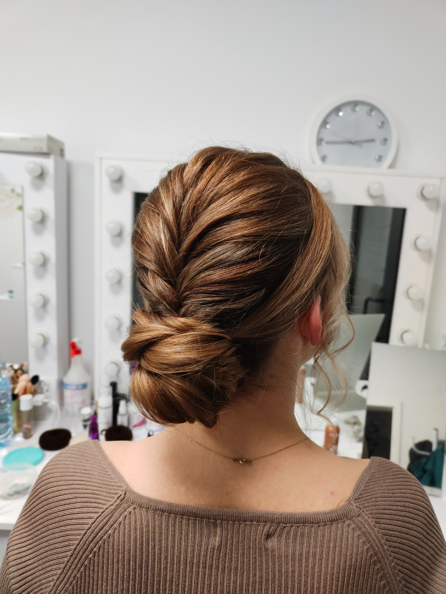 Upięcie włosów w kok z warkoczem, widok z tyłu, na tle toaletki z lustrem i oświetleniem.