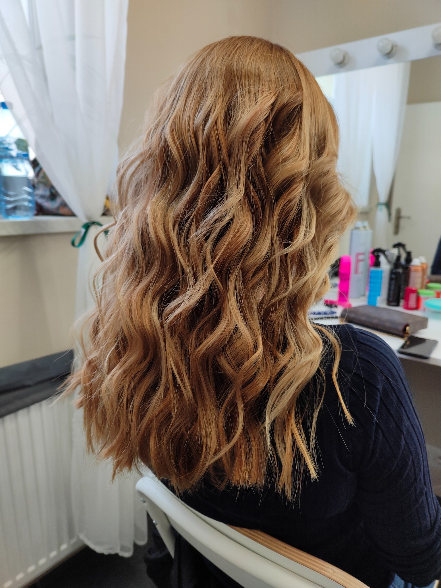 Długie, falowane włosy w odcieniach blondu i miedzi, widoczne od tyłu, w tle lustro z oświetleniem i kosmetyki na blacie.