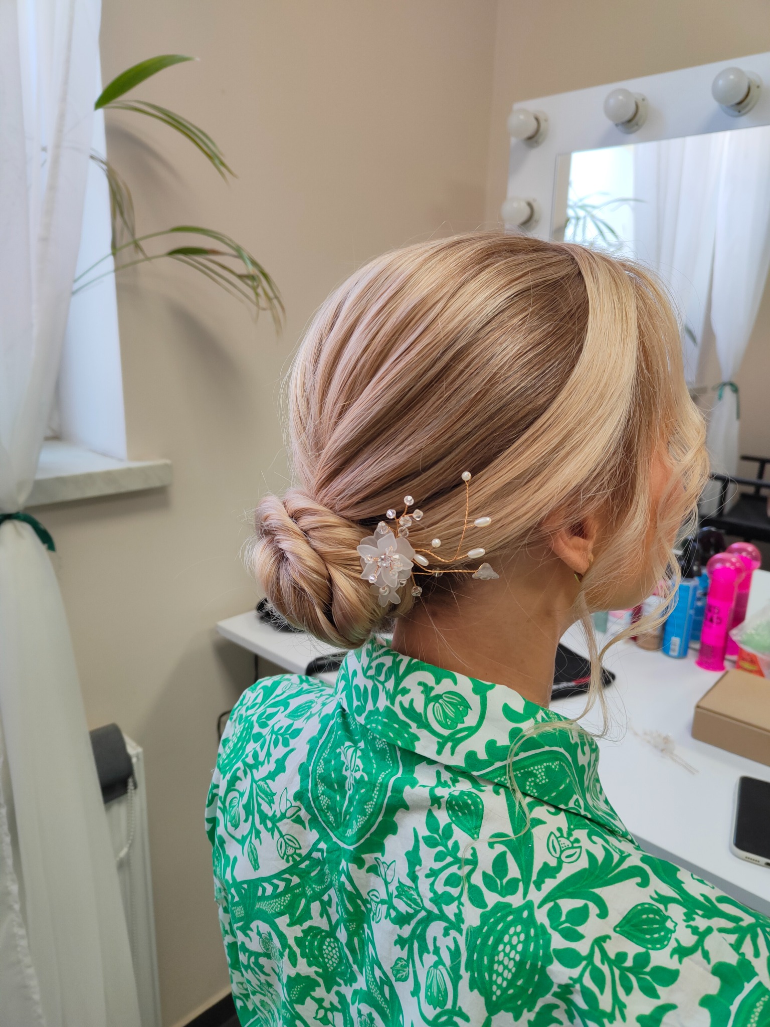 Upięcie blond włosów w niski kok z dekoracyjną spinką z perłami i kwiatem, widok z boku, w tle lustro z oświetleniem i kosmetyki