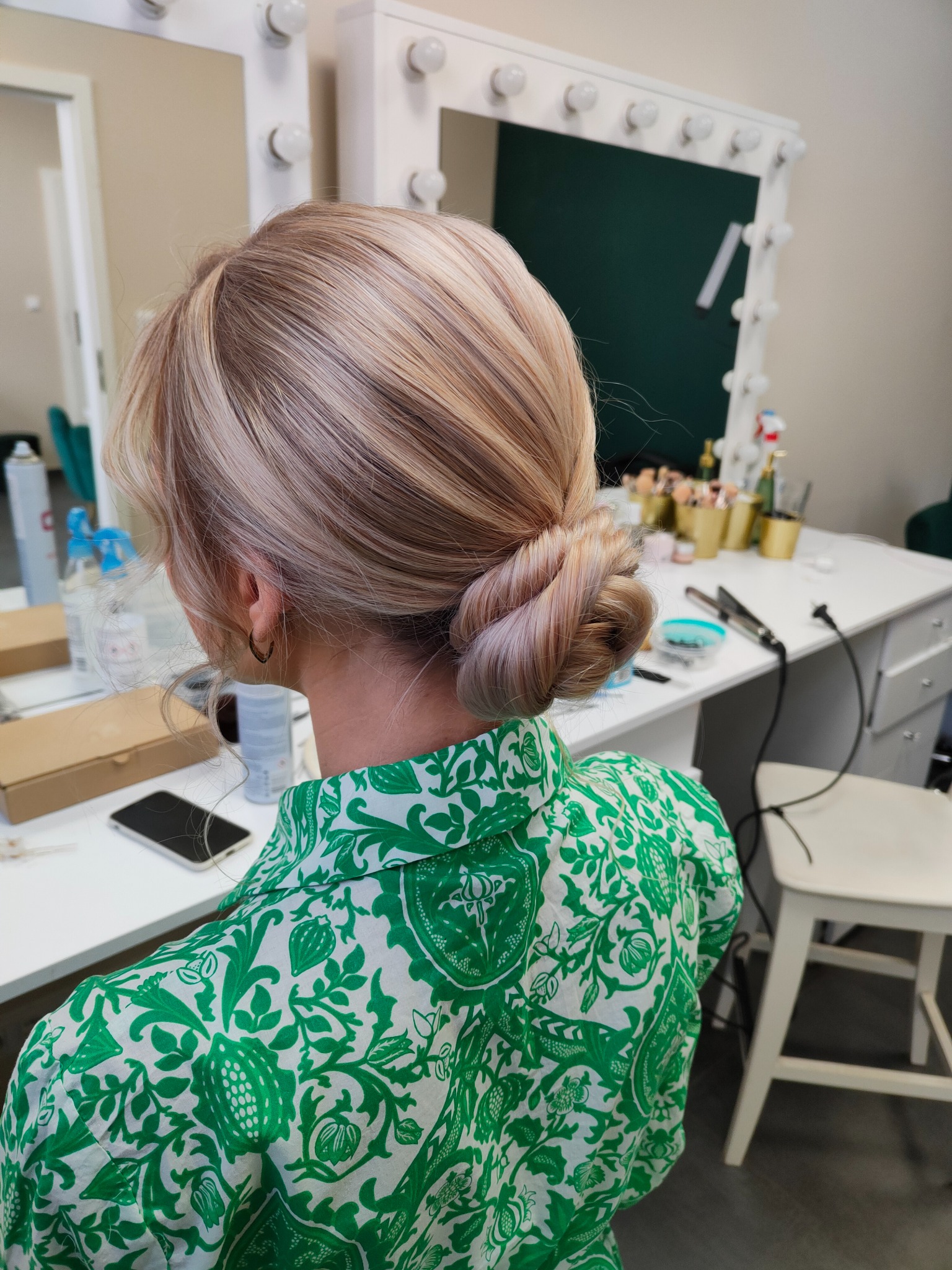 Upięcie blond włosów w kok z tyłu głowy, widoczny fragment zielonej koszuli w biały wzór roślinny, w tle toaletka z lustrem i akcesoriami.
