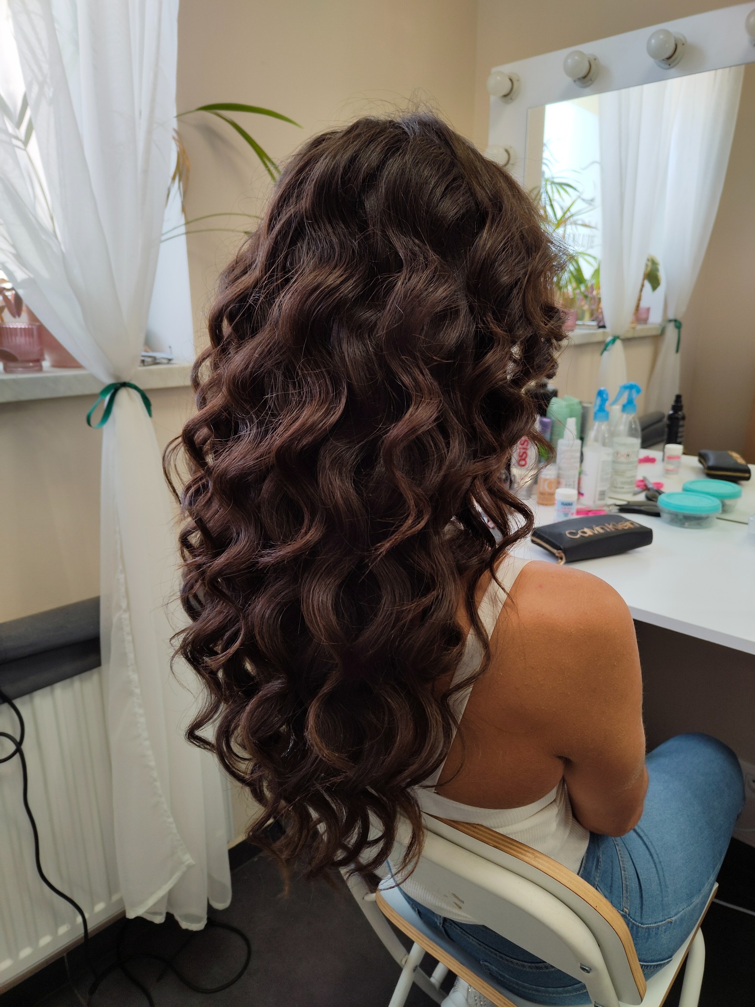 Brunetka z długimi, brązowymi, kręconymi włosami, siedząca tyłem do kamery w salonie, widoczne akcesoria do stylizacji na białym stole z oświetleniem.