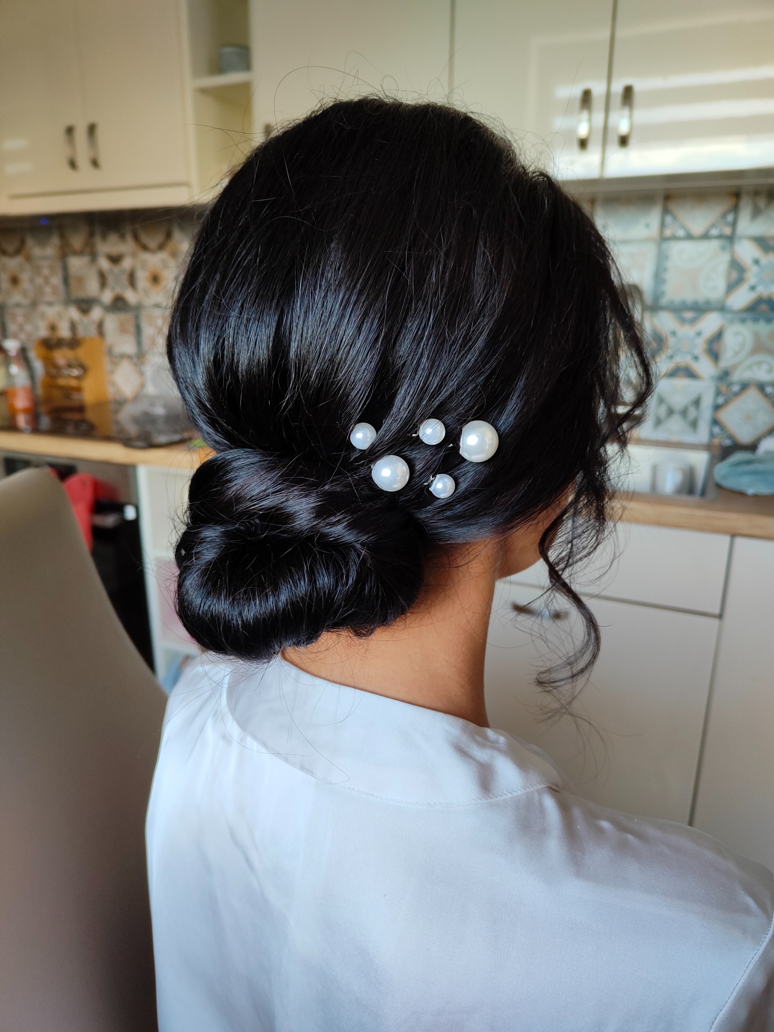 Elegancka fryzura upięta w kok z boku głowy, ozdobiona czterema wsuwkami z perłami, na tle kuchni.