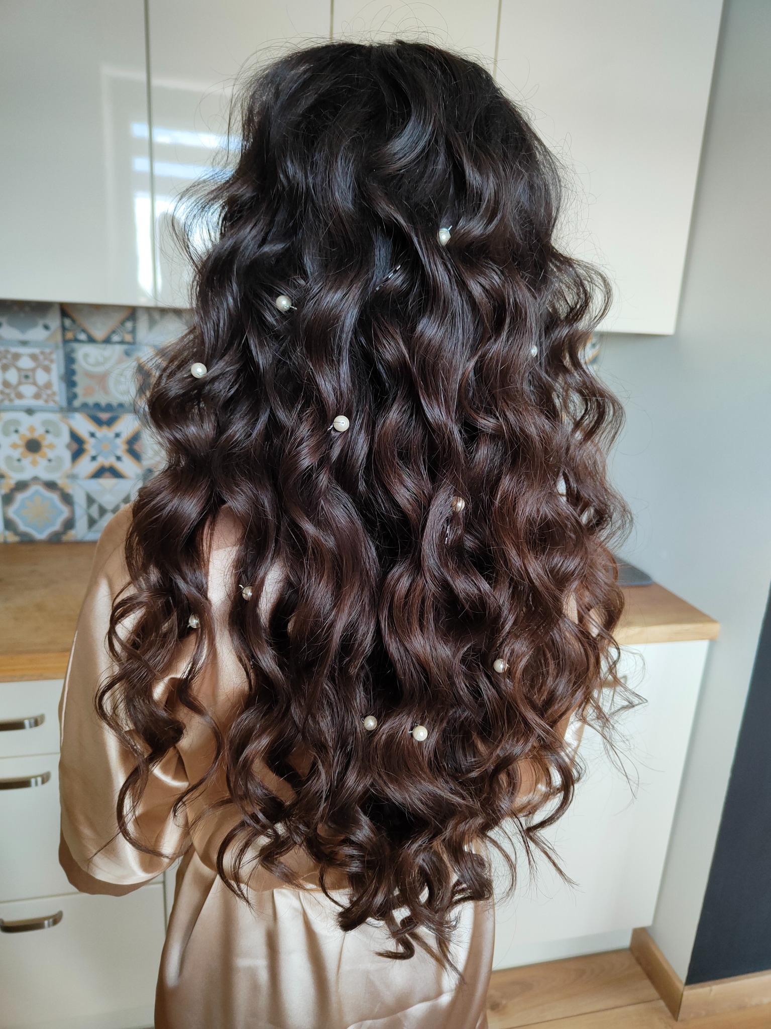 Długie, ciemne, kręcone włosy upięte z tyłu głowy, ozdobione małymi perłami na wsuwkach, widok z tyłu, w tle fragment kuchni.