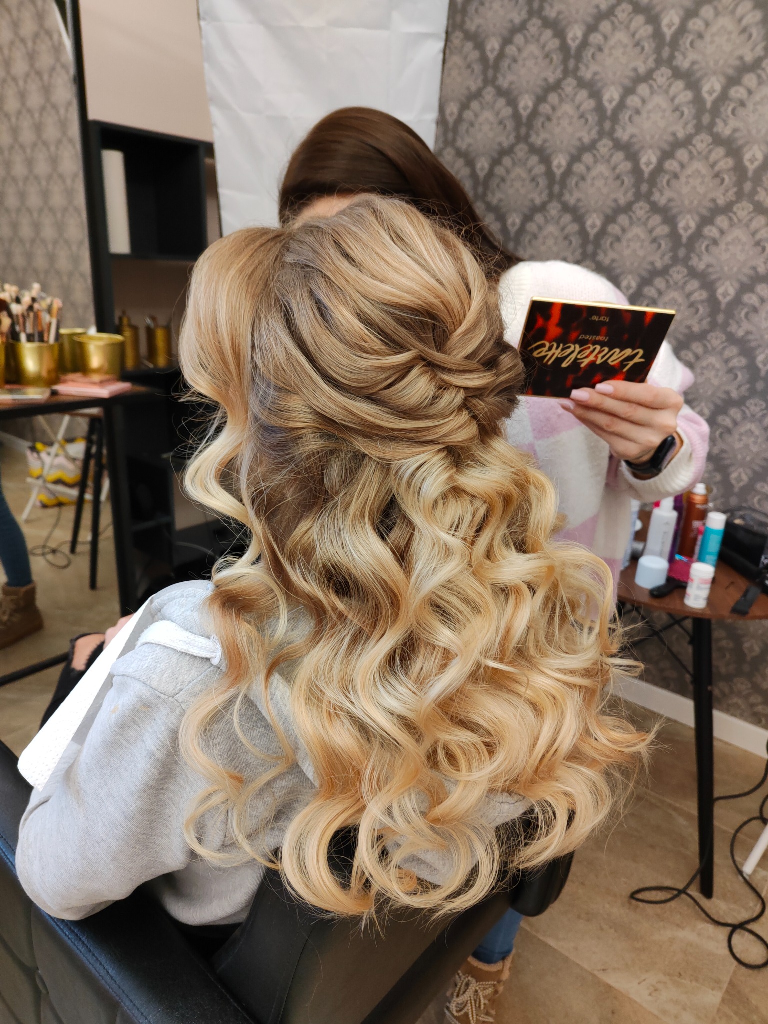 Upięcie blond włosów z lokami, widok z tyłu, stylistka trzyma paletę cieni Tartelette Toasted w dłoni.