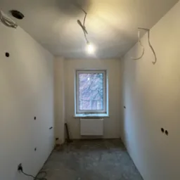 Pokój w trakcie remontu: gołe ściany z otworami na gniazdka, betonowa podłoga, okno z grzejnikiem, wiszące przewody elektryczne i surowa żarówka.