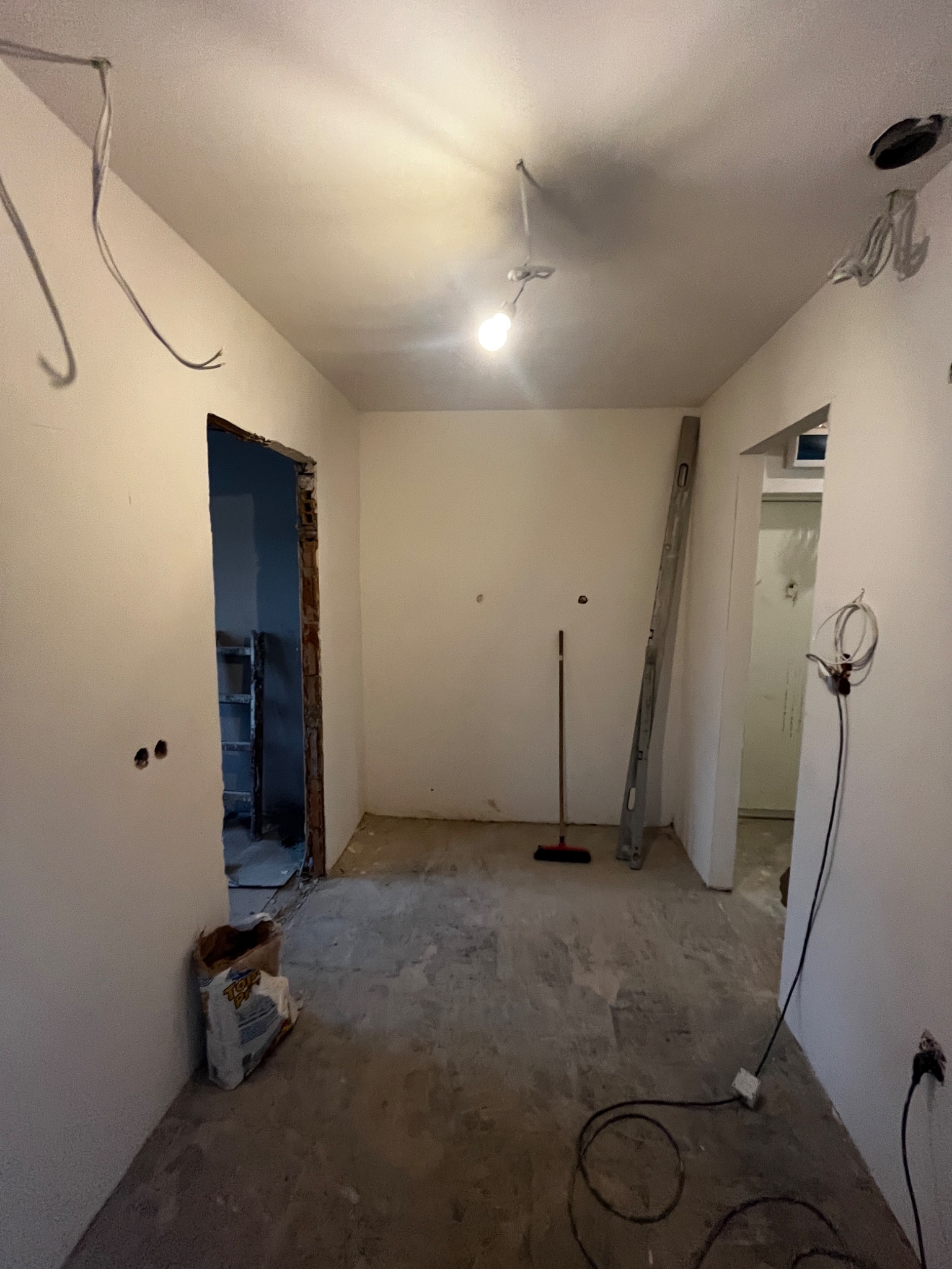Pomieszczenie w trakcie remontu: gołe ściany, przewody wystające z sufitu, worek z zaprawą przy ścianie, drabina oparta o ścianę, widoczny brak wykończenia ościeżnicy.