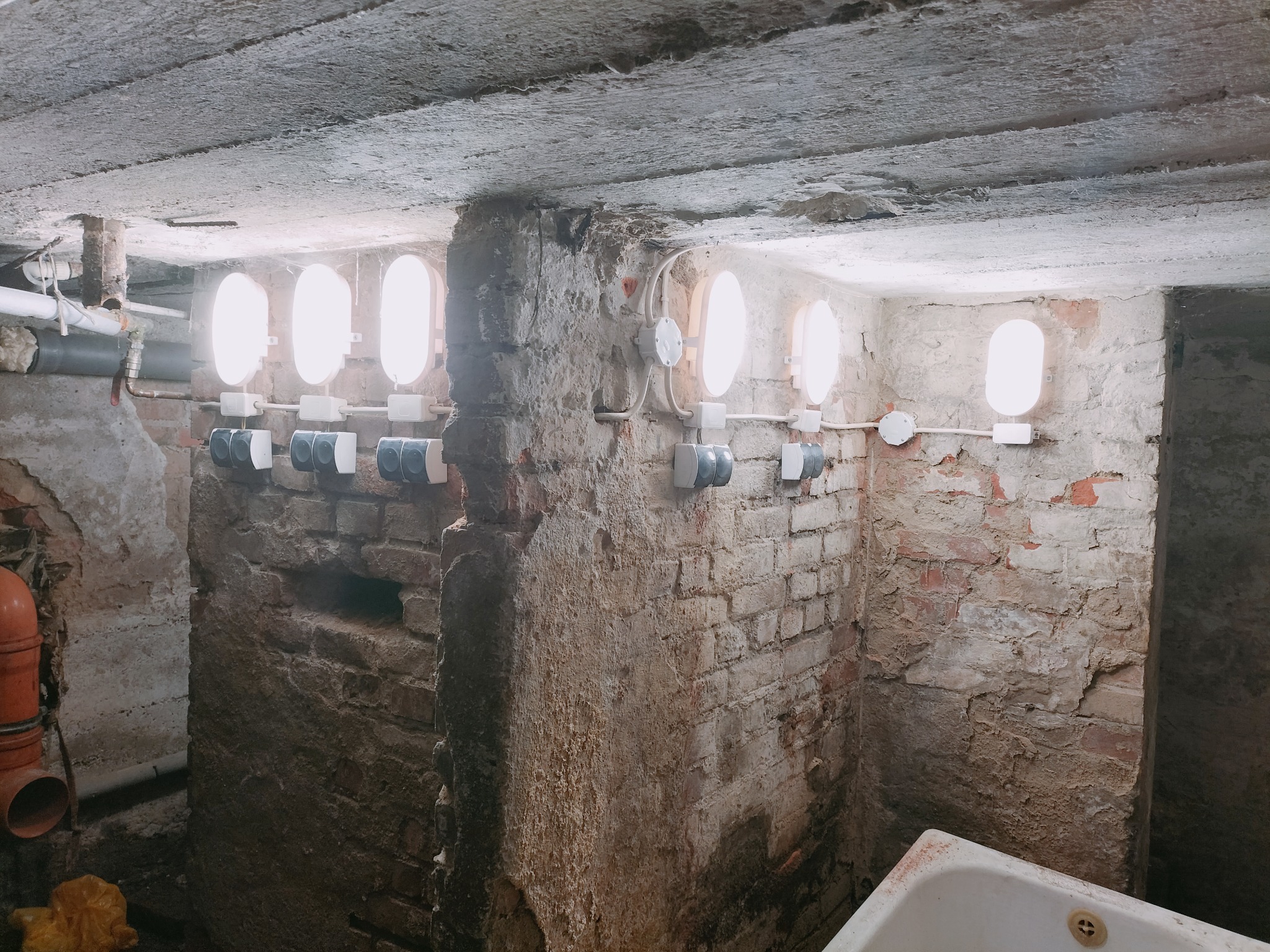 Piwnica z surowymi ceglanymi ścianami i betonowym sufitem, oświetlona rzędem owalnych lamp naściennych z szarymi przełącznikami, widoczne rury instalacyjne.