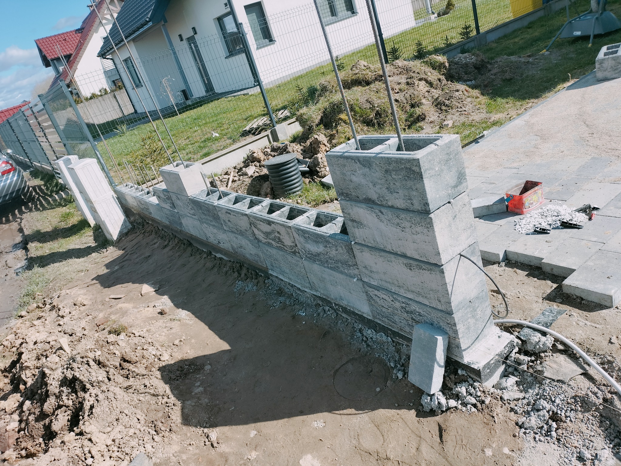 Wznoszenie ogrodzenia z szarego bloczka betonowego, widoczne pręty zbrojeniowe, teren wokół w trakcie prac ziemnych, częściowo ułożona kostka brukowa, dom w tle.