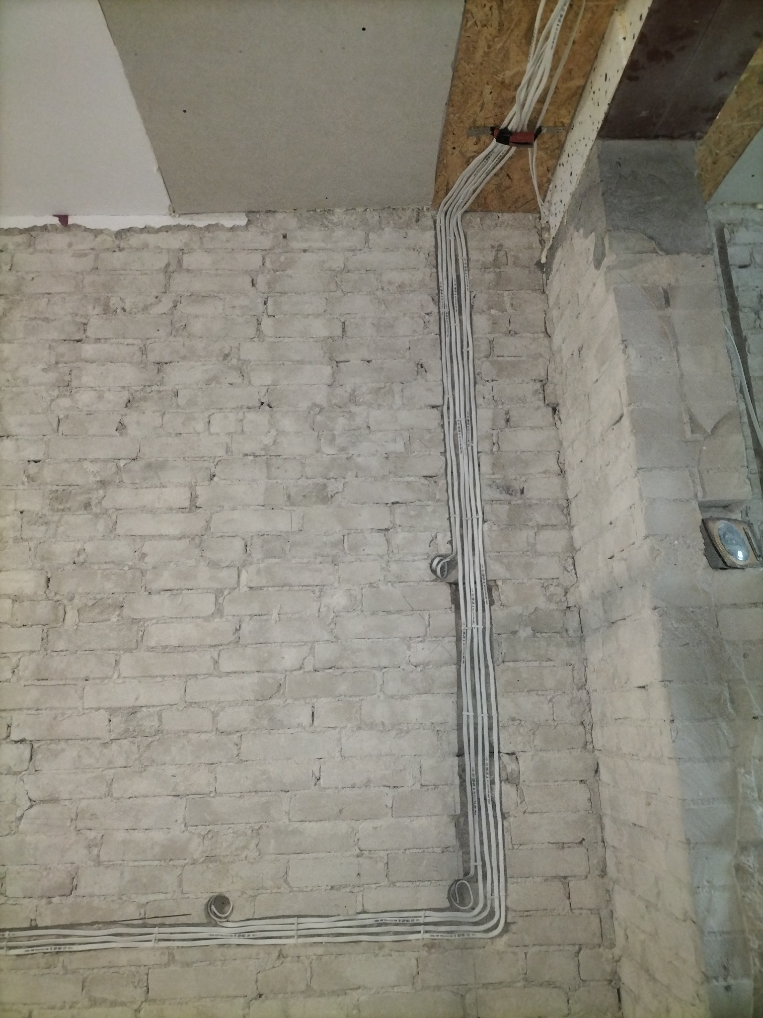 Białe przewody elektryczne ułożone pionowo na surowej, pomalowanej na biało ścianie z cegły, z widocznym fragmentem sufitu z płyty OSB