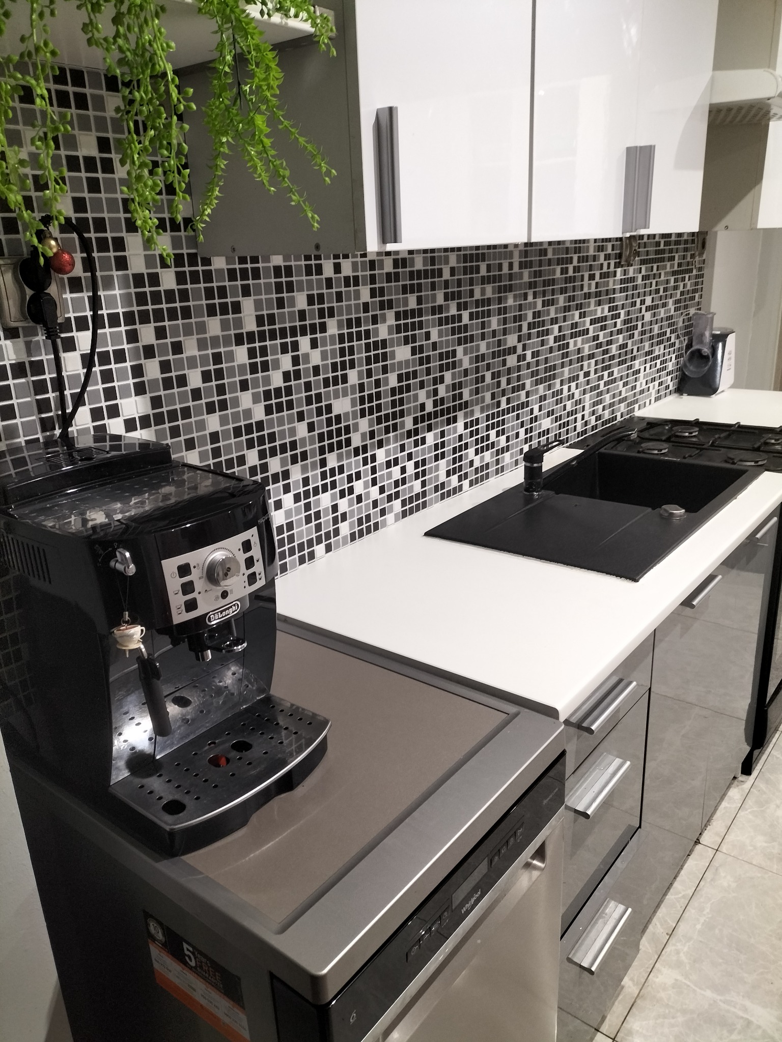 Nowoczesna kuchnia z czarno-białą mozaiką na ścianie, białym blatem, czarnym zlewem i płytą gazową, z wiszącą rośliną i ekspresem do kawy DeLonghi z zawieszką w kształcie filiżanki.
