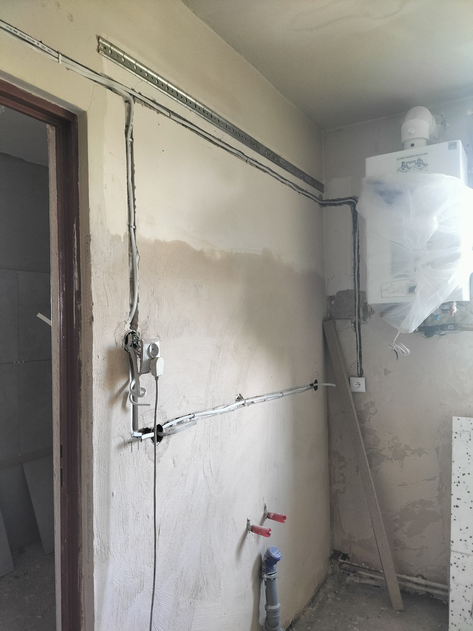 Wnętrze w trakcie remontu z widocznymi przewodami elektrycznymi w korytach, puszką podtynkową z wystającymi kablami, instalacją hydrauliczną i zabezpieczonym folią piecem gazowym na ścianie.