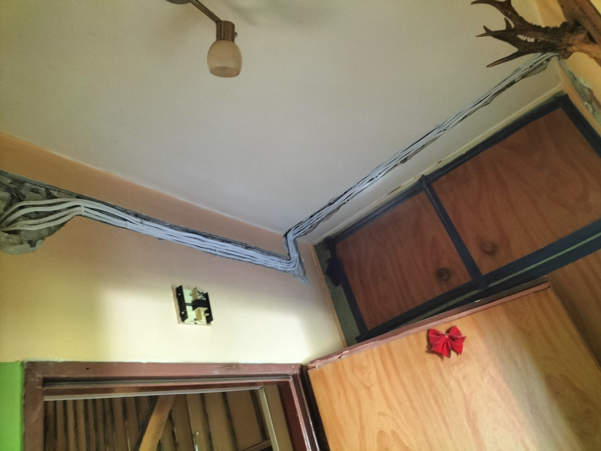 Kable elektryczne ułożone wzdłuż sufitu i ściany, widoczna puszka elektryczna bez osprzętu, drewniane szafki z czerwoną kokardką dekoracyjną, fragment poroża jelenia.