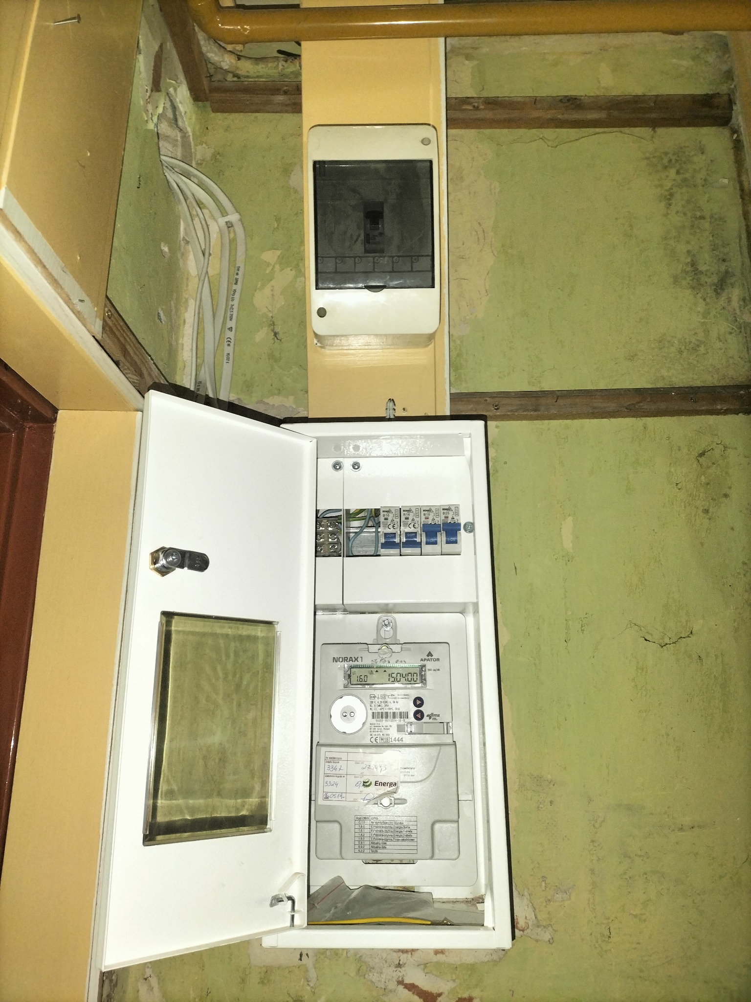 Otwarta skrzynka elektryczna z licznikiem i bezpiecznikami na tle zielonej ściany z widocznymi kablami w ścianie, z bliska.