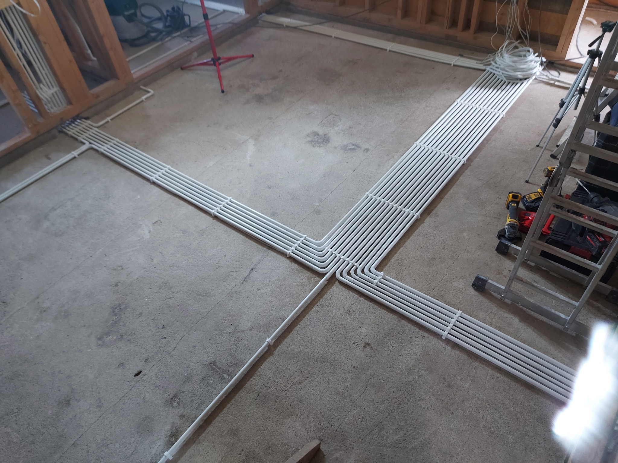 Białe rury prowadzone po betonowej podłodze w surowym stanie, tworzące krzyżujące się wiązki, z widoczną drabiną i narzędziami w tle.