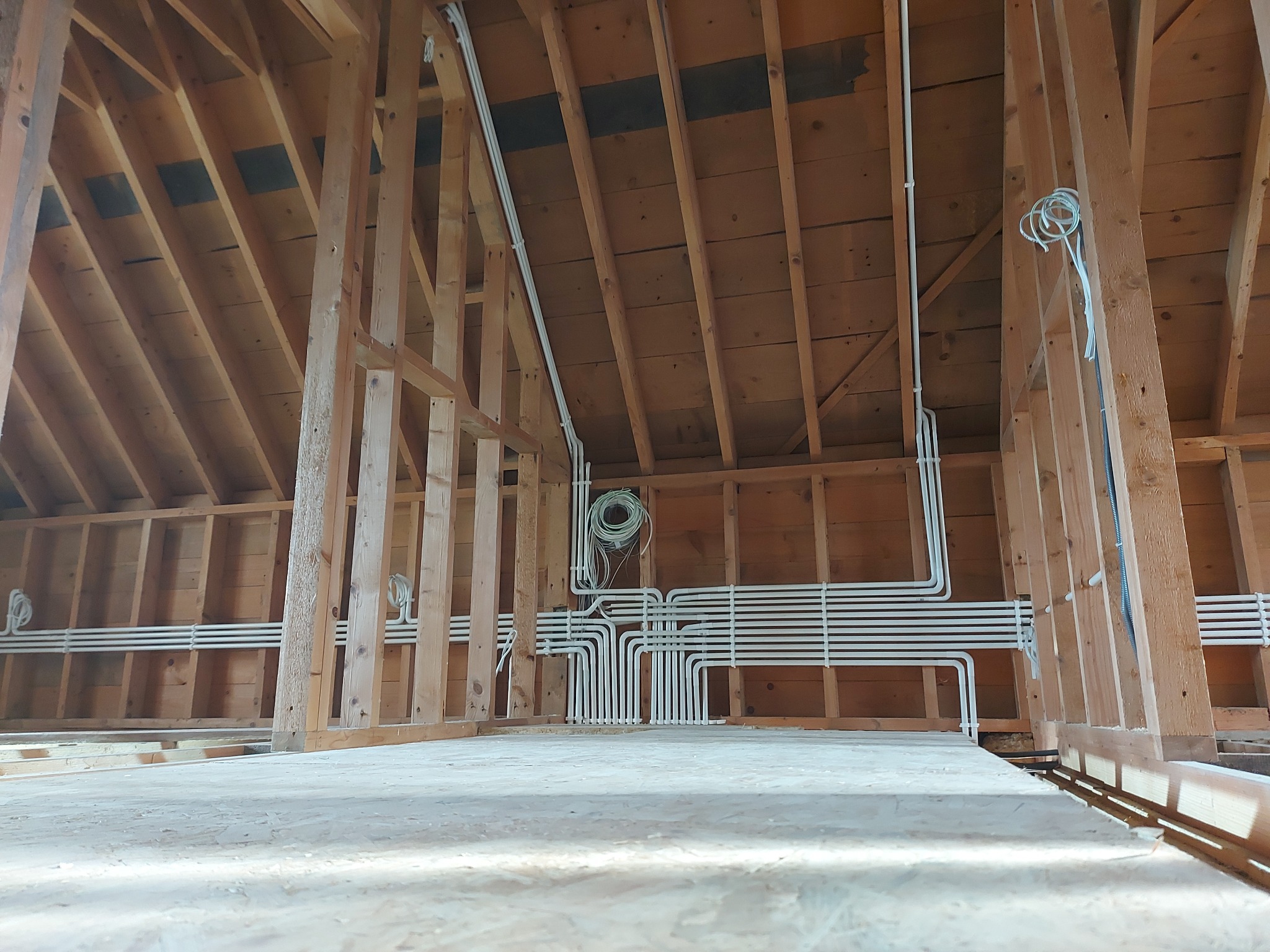 Białe rury instalacji elektrycznej ułożone równolegle w drewnianej konstrukcji poddasza w trakcie budowy, widok z dołu.