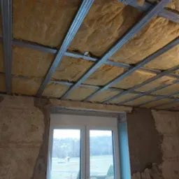 Montaż izolacji z wełny mineralnej w metalowym stelażu sufitu podwieszanego na poddaszu, widok na okno balkonowe i krajobraz za nim.