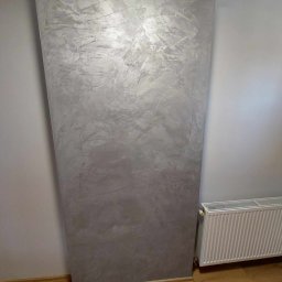 Haustechnik REMONTY I WYKOŃCZENIA Andrzej Dychus - Dekoracyjna ściana w odcieniach szarości z efektem przetarć, przylegająca do grzejnika i jasnej ściany, na drewnianej podłodze.
