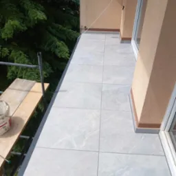 Jasnoszare płytki gresowe imitujące marmur na balkonie, widoczny fragment rusztowania z wiadrem farby, zieleń drzew w tle.