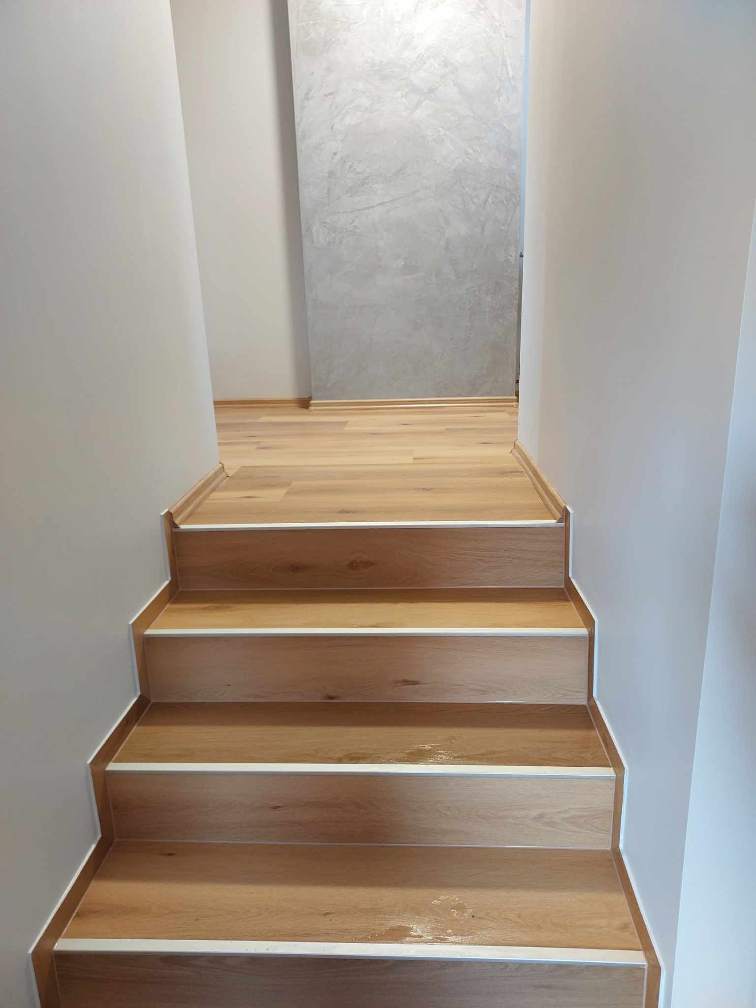 Schody wyłożone panelami drewnopodobnymi z białymi listwami przypodłogowymi, widok z dołu na górę, ściany pomalowane na jasny kolor, w tle ściana z efektem dekoracyjnym betonu.