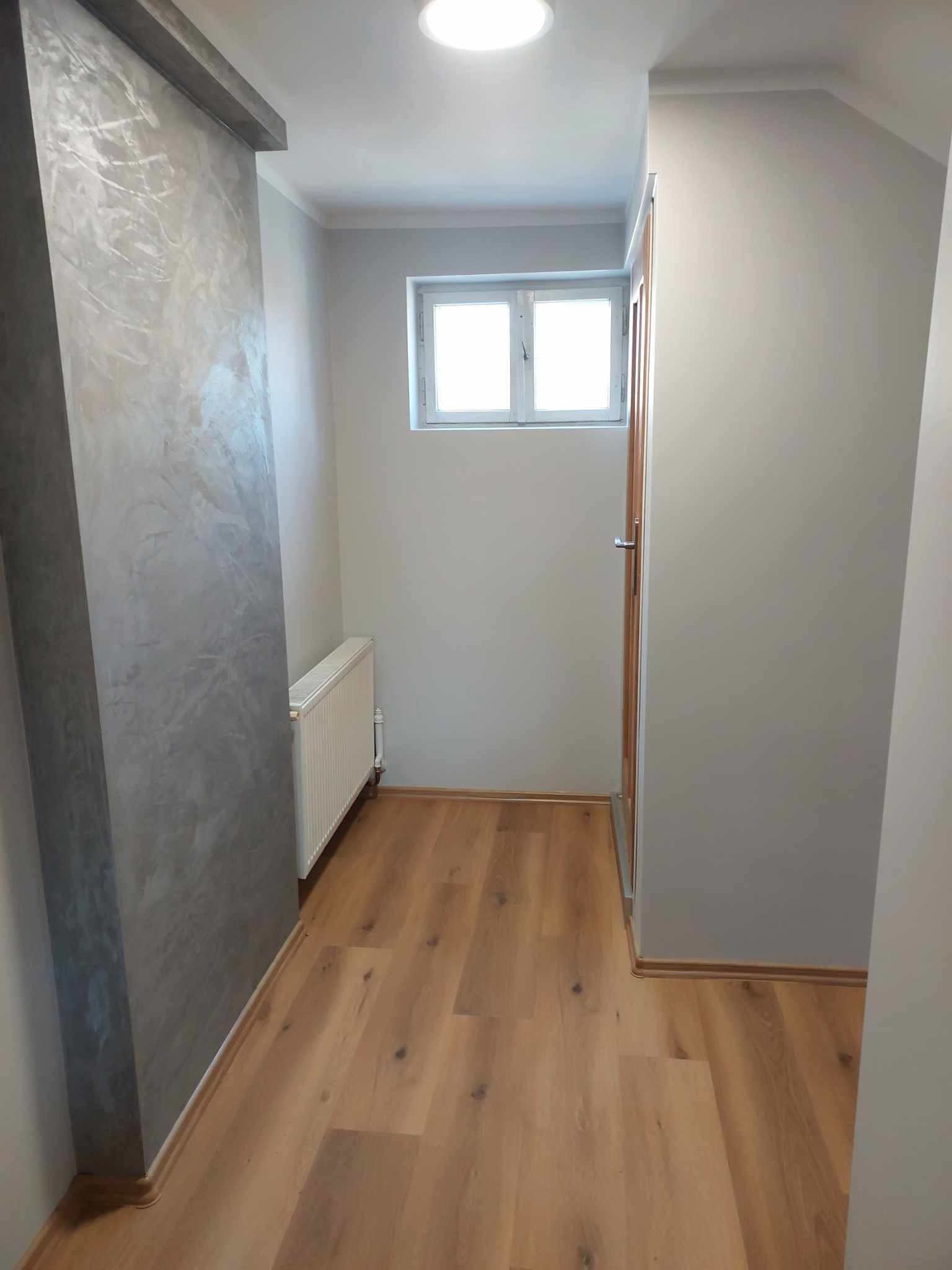 Wąski korytarz z dekoracyjną ścianą w odcieniach szarości, panelami podłogowymi w kolorze jasnego drewna, białym grzejnikiem i oknem z białymi ramami.