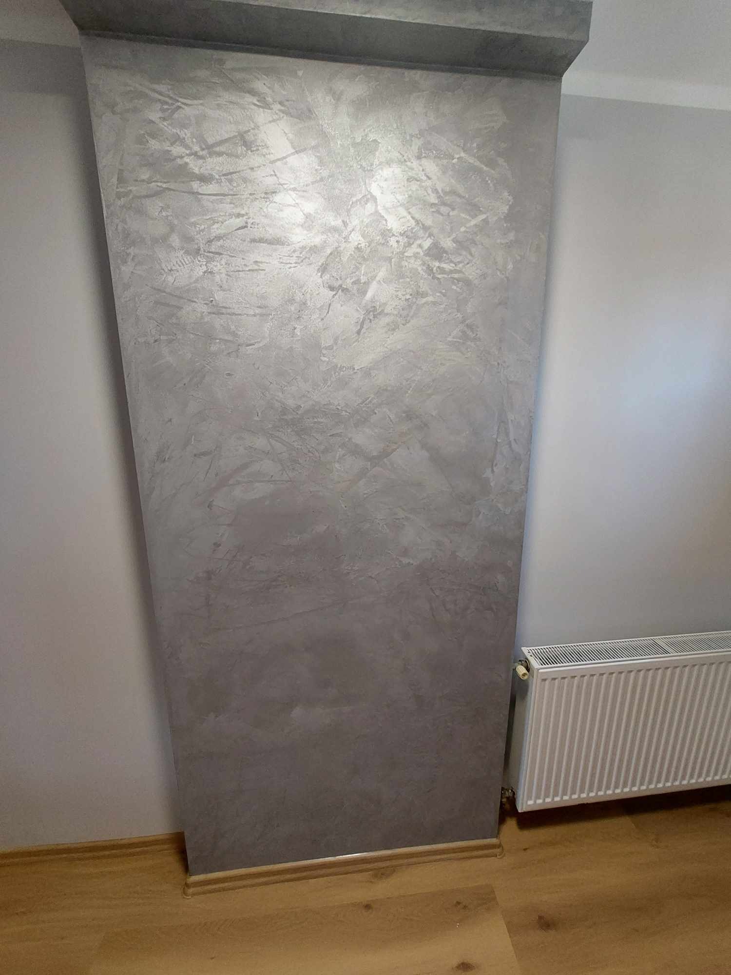 Dekoracyjna ściana w odcieniach szarości z efektem przetarć, przylegająca do grzejnika i jasnej ściany, na drewnianej podłodze.