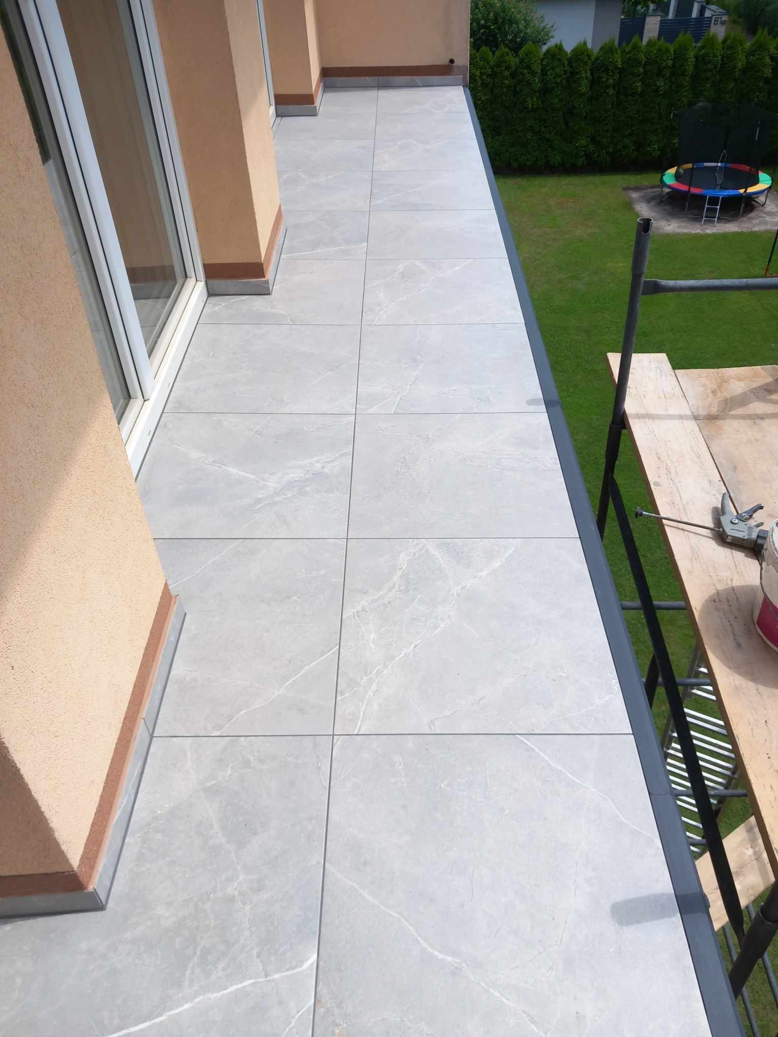 Wykończony balkon z szarymi płytkami imitującymi marmur, widok z góry, z fragmentem rusztowania i narzędzi na tle zielonego trawnika.