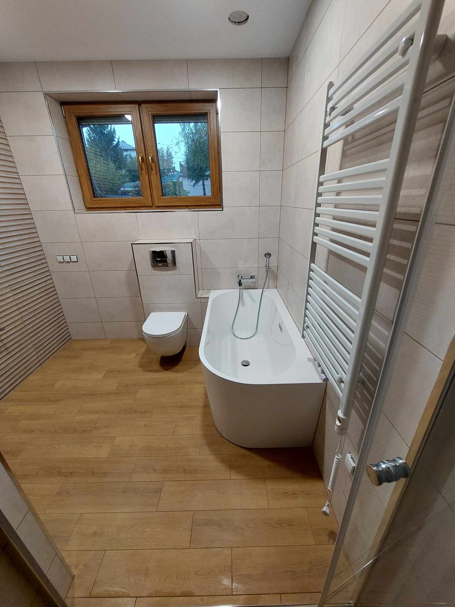 Nowoczesna łazienka z beżowymi płytkami, drewnopodobną podłogą, białą wanną asymetryczną i grzejnikiem drabinkowym; widok z perspektywy wejścia.