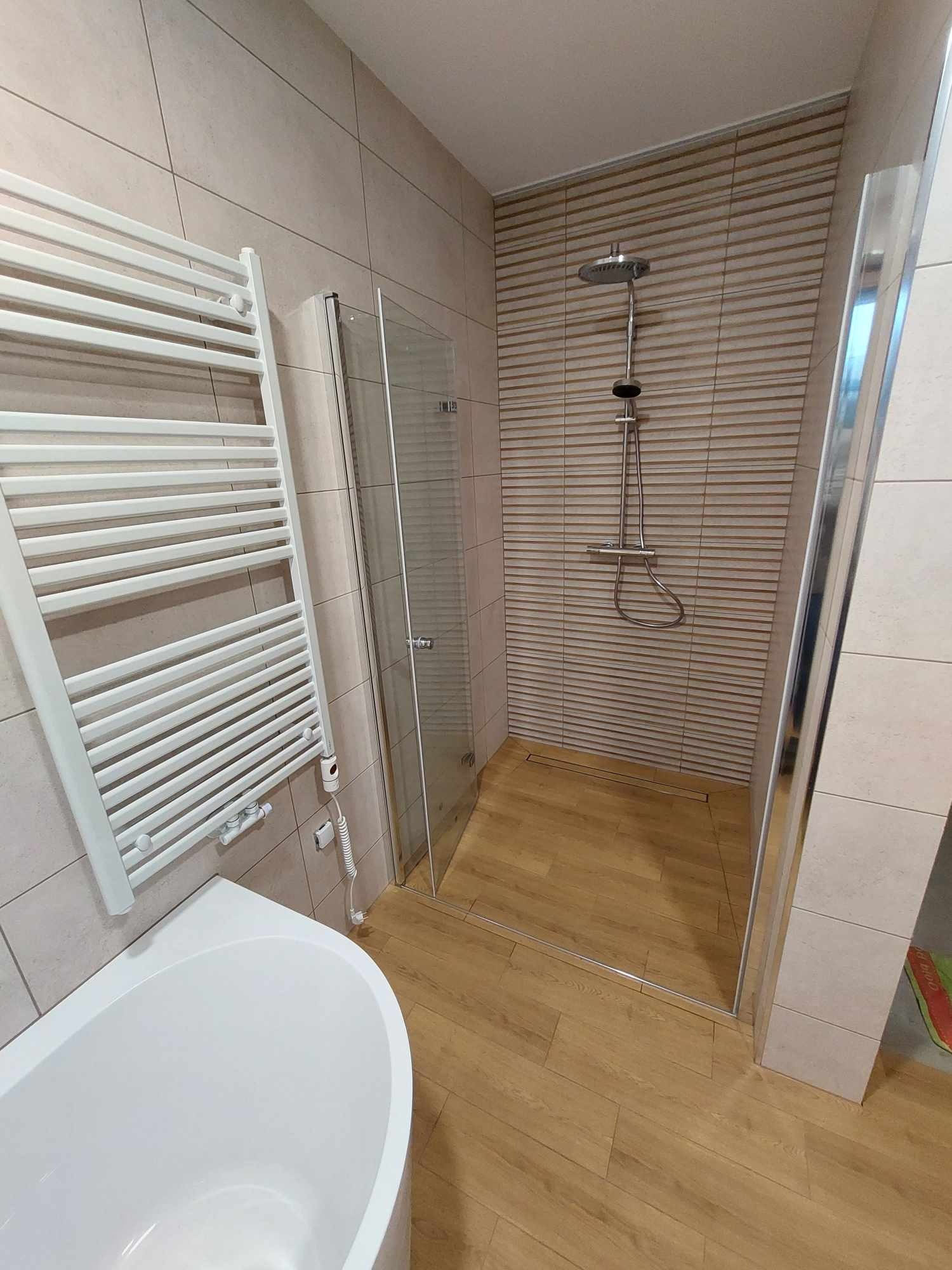 Nowoczesna łazienka z kabiną prysznicową typu walk-in, grzejnikiem drabinkowym i wanną, wykończona płytkami w odcieniach beżu i brązu z drewnopodobną podłogą i odpływem liniowym