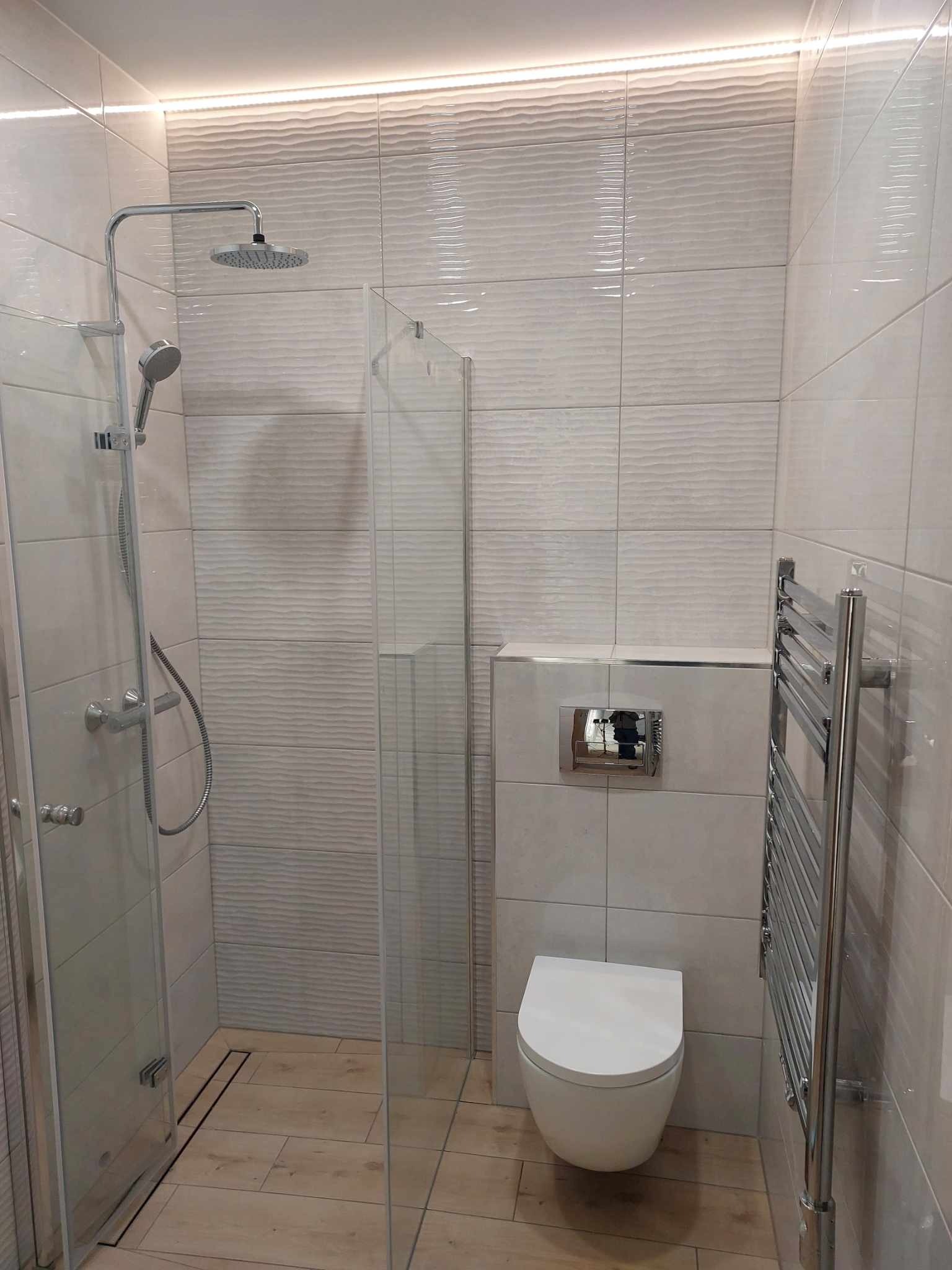 Nowoczesna łazienka z kabiną prysznicową typu walk-in, podwieszaną miską WC, grzejnikiem drabinkowym i płytkami z subtelnym falistym wzorem oraz oświetleniem LED podsufitowym.