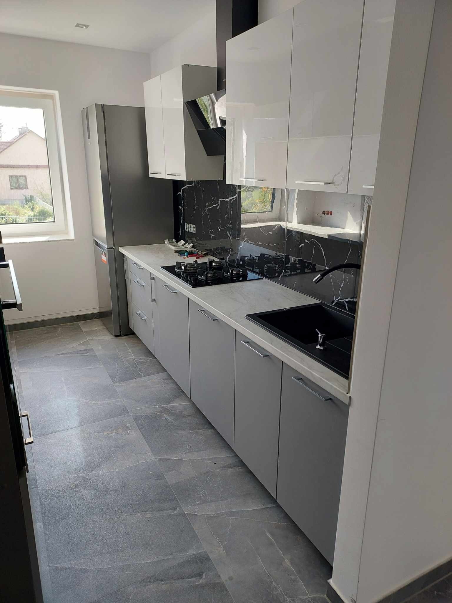 Nowoczesna kuchnia z szarymi szafkami, białymi górnymi szafkami, czarnym blatem imitującym marmur i szarą podłogą z płytek imitujących kamień.