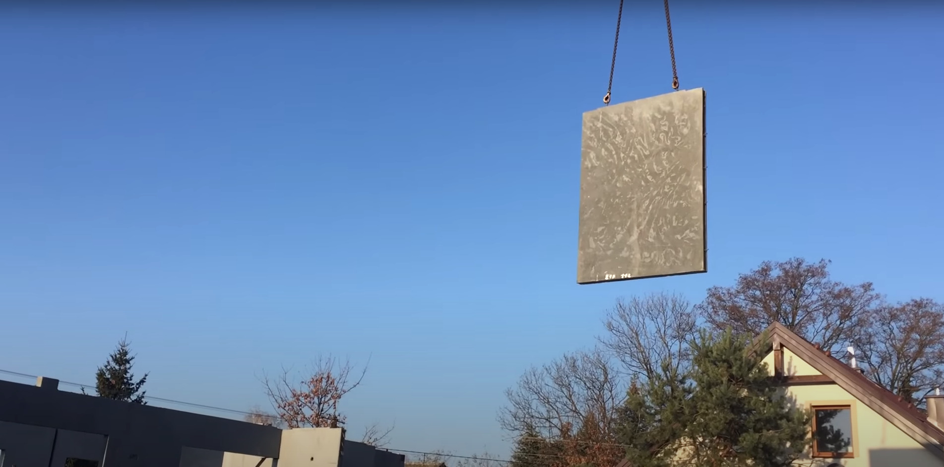 Panel z betonu architektonicznego zawieszony na łańcuchach dźwigu na tle błękitnego nieba i zabudowy jednorodzinnej.