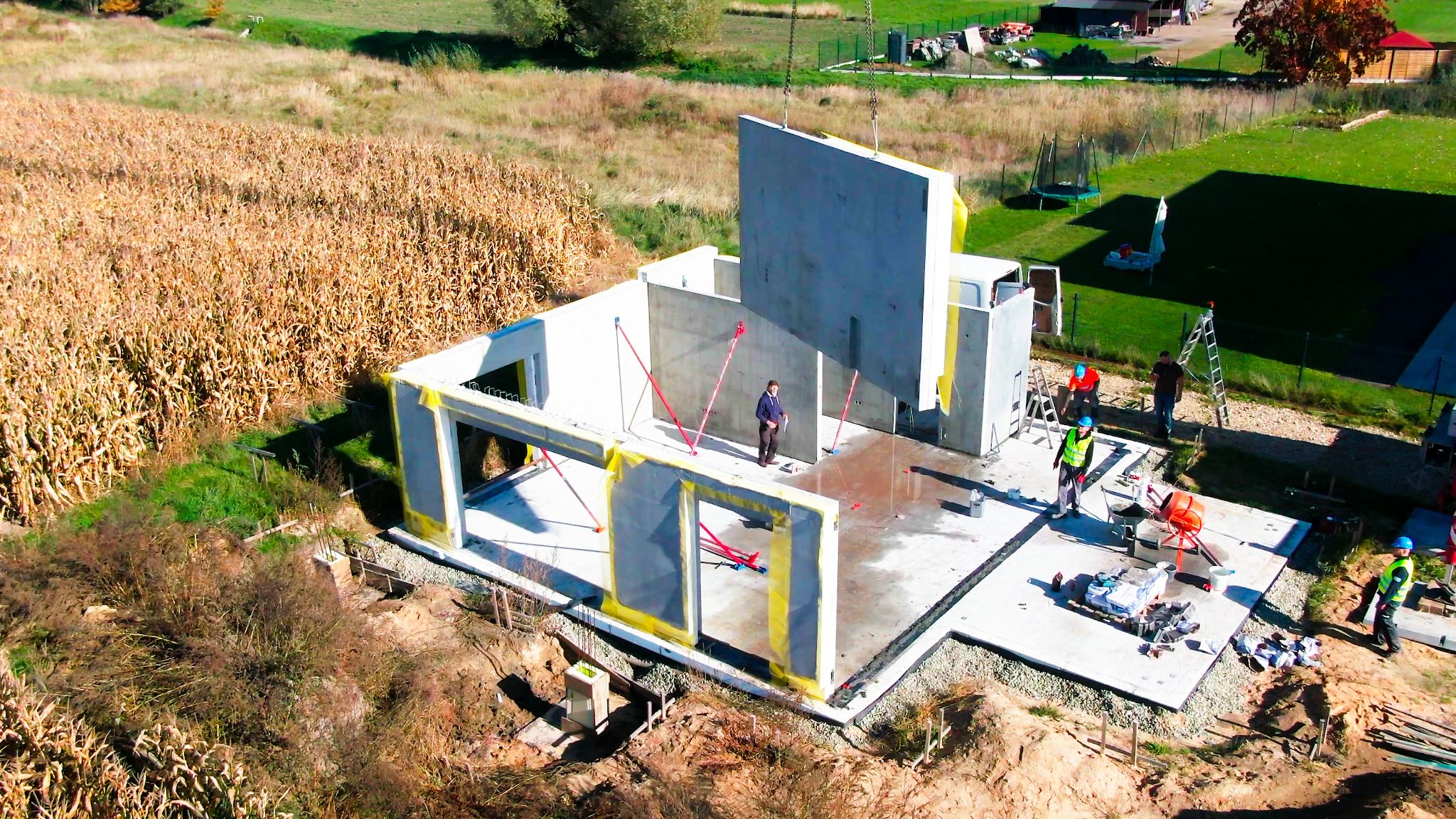 Montaż prefabrykowanych ścian na budowie domu, widok z góry na teren z polem kukurydzy w tle.