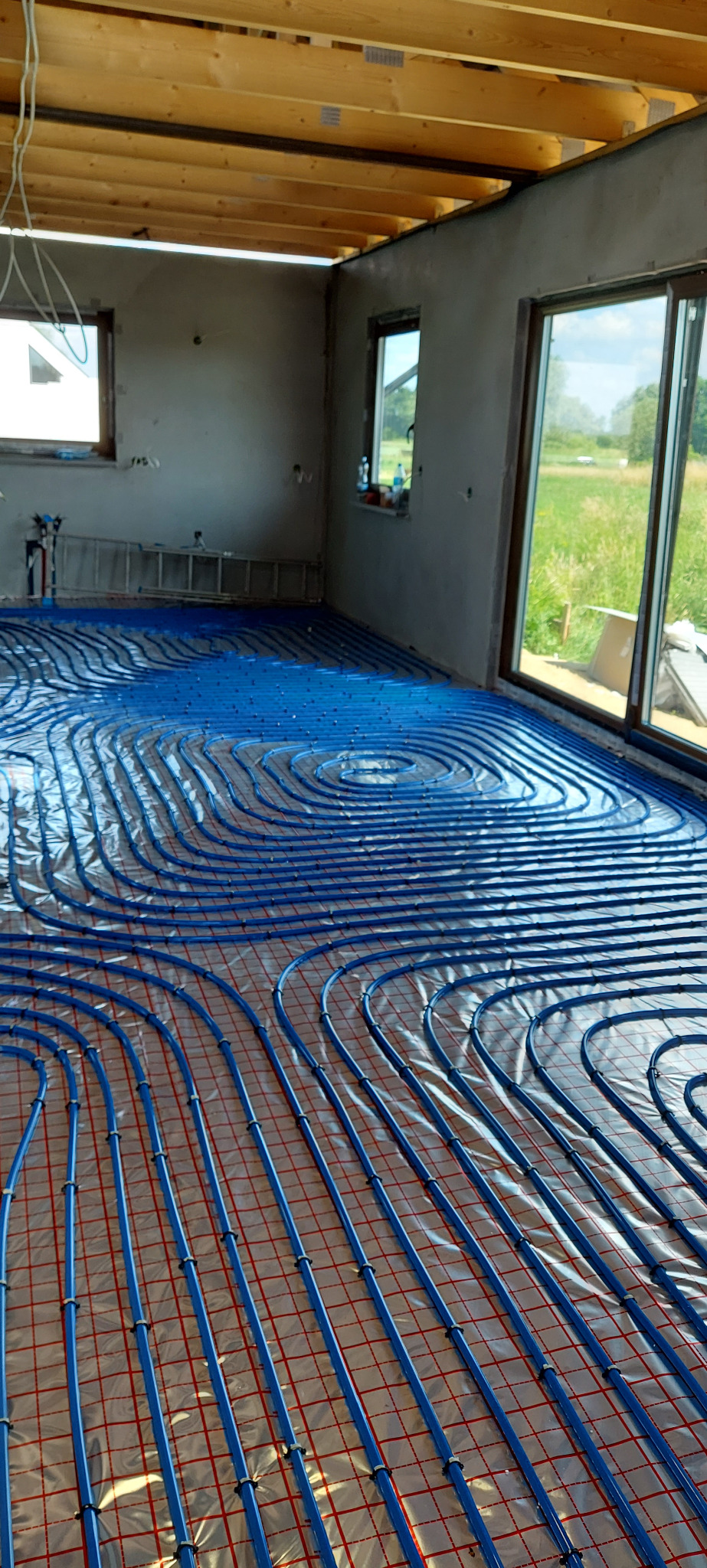 Instalacja ogrzewania podłogowego z niebieskich rur na folii izolacyjnej w surowym wnętrzu domu w trakcie budowy, widoczne okna i drewniana konstrukcja dachu.