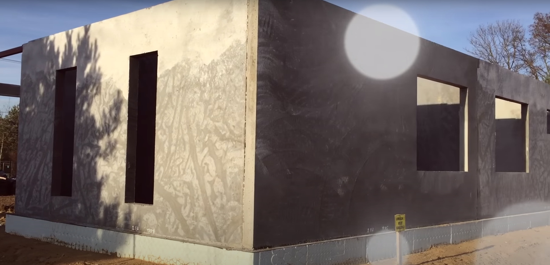 Surowy, betonowy budynek w trakcie budowy, z widocznymi otworami okiennymi i fundamentami, oświetlony jasnym światłem słonecznym, z żółtą tabliczką ostrzegawczą w pobliżu.