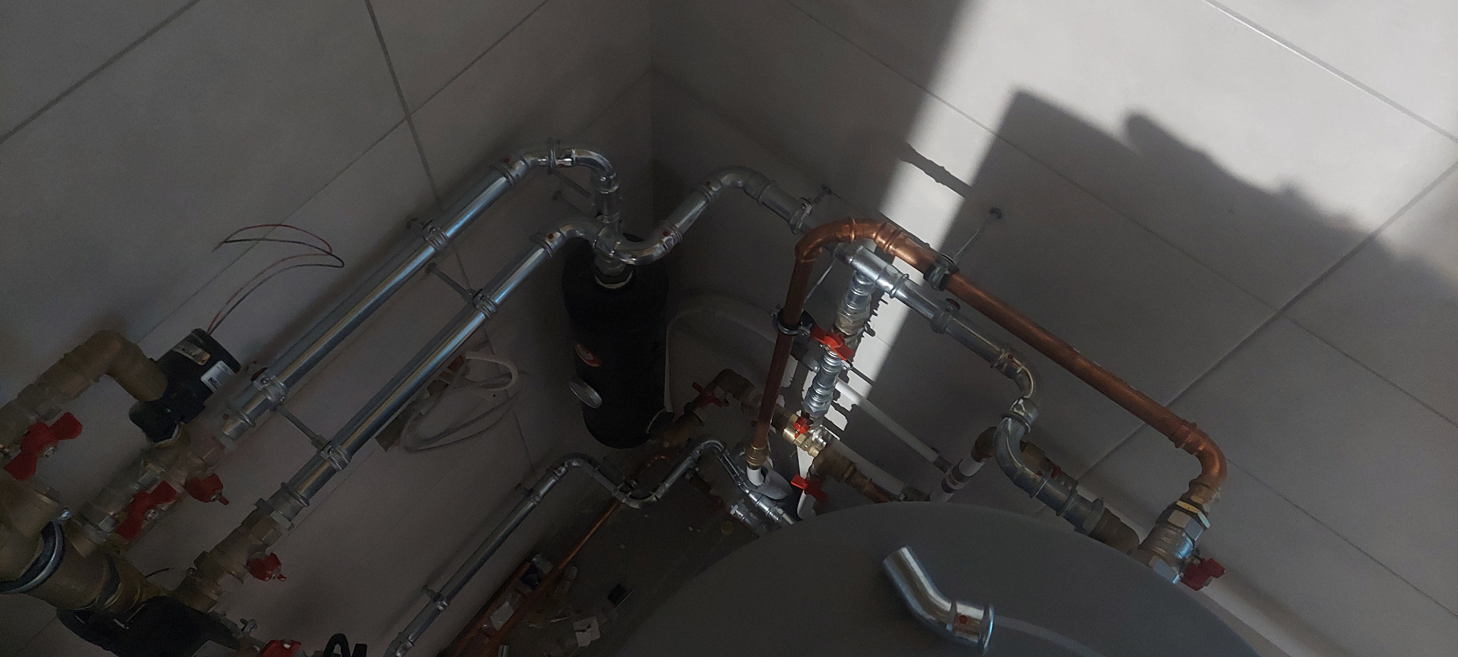 Złożona instalacja grzewcza z rur miedzianych i ocynkowanych, widoczna z perspektywy sufitu, z zaworami z czerwonymi uchwytami i czarnym zbiornikiem wyrównawczym.