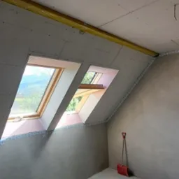 Poddasze z oknami dachowymi w trakcie wykańczania, widoczne płyty gipsowo-kartonowe, ocieplenie wełną mineralną i czerwona łopata oparta o ścianę.