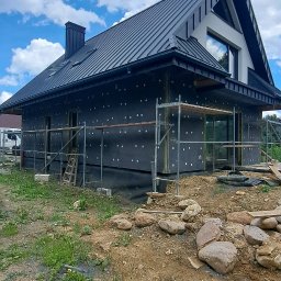 Damian Siwek - Dom jednorodzinny w trakcie ocieplania, widoczne rusztowania, czarna membrana z białymi kropkami na elewacji, ciemny dach z blachy na tle błękitnego nieba z chmurami.