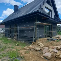 Dom jednorodzinny w trakcie ocieplania, widoczne rusztowania, czarna membrana z białymi kropkami na elewacji, ciemny dach z blachy na tle błękitnego nieba z chmurami.