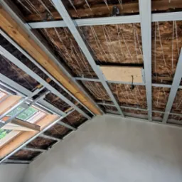 Poddasze w trakcie ocieplania: widoczne wełna mineralna, metalowy stelaż, sznurki podtrzymujące izolację oraz okno dachowe, ściany częściowo wykończone gładzią, na podłodze czarny worek na śmieci.