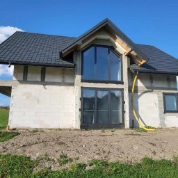 Damian Siwek - Dom w trakcie budowy z szarym dachem i dużymi oknami. Ściany z białych bloczków, widoczne elementy konstrukcyjne. Żółty wąż przy ścianie. Błękitne niebo w tle.