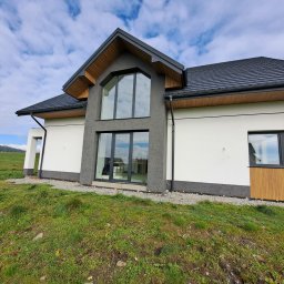 Damian Siwek - Nowoczesny dom jednorodzinny z dużymi oknami i elewacją w odcieniach bieli i szarości, otoczony zielonym trawnikiem i błękitnym niebem z chmurami.